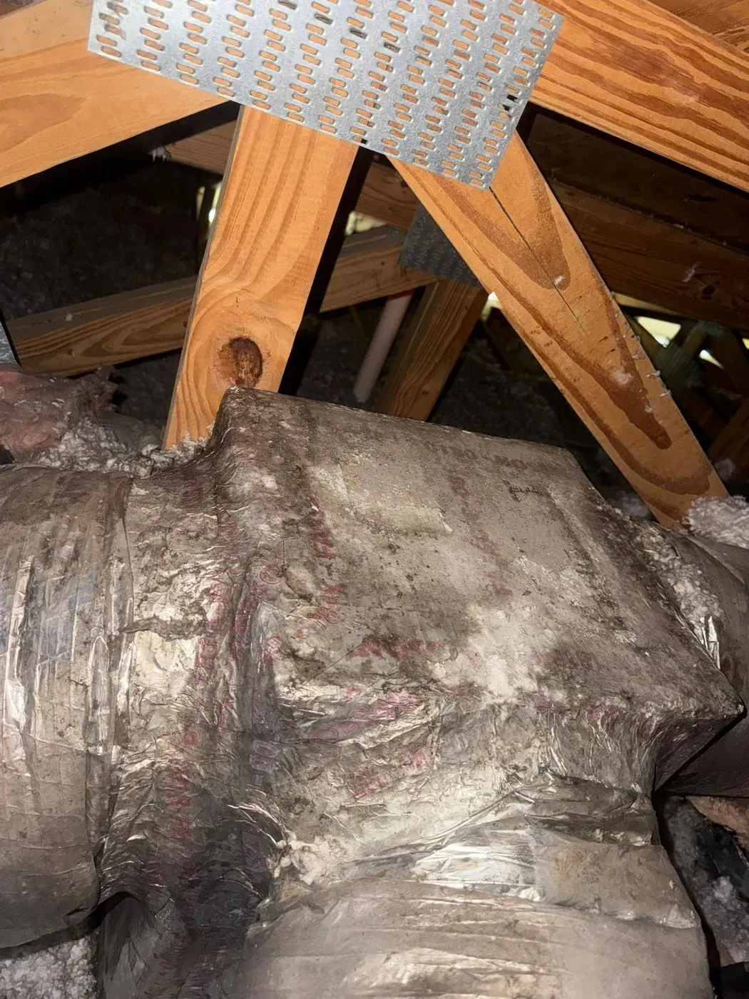 Dust-caked insulated duct runs between wood roof trusses in an attic