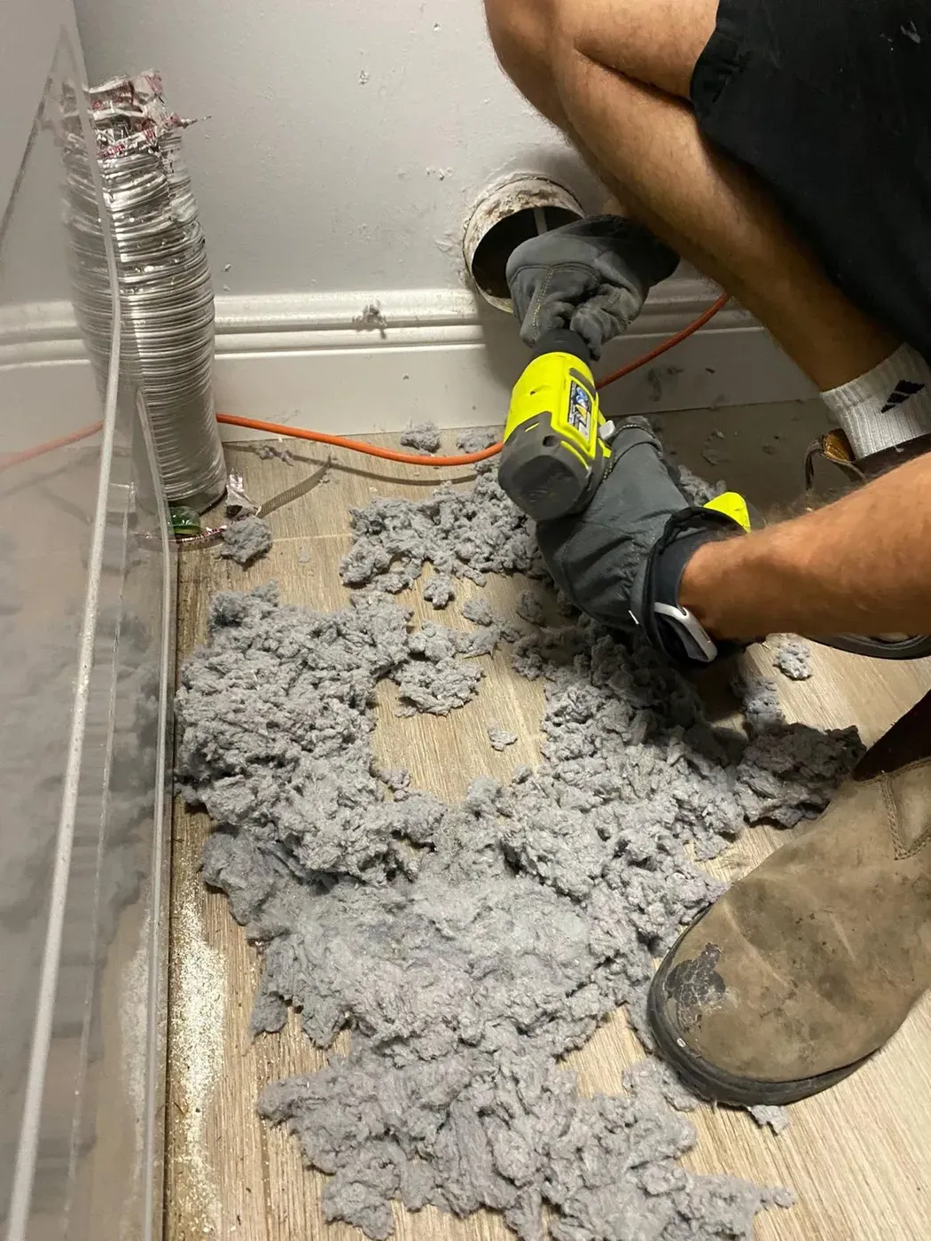 Technician using a cordless rotary tool at a wall dryer vent with a pile of removed gray lint on the floor