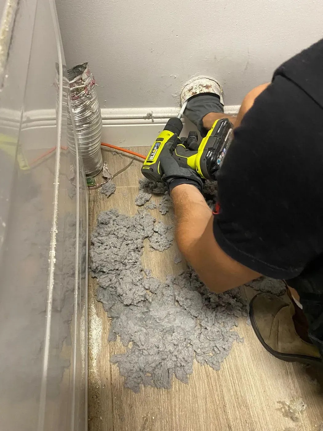 Technician kneeling to clean a wall dryer vent, with removed lint and the transition duct on the floor