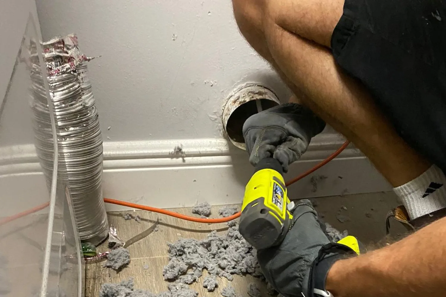 Technician using a cordless rotary tool at a wall dryer vent stub, with a large pile of gray lint pulled from the duct on the floor